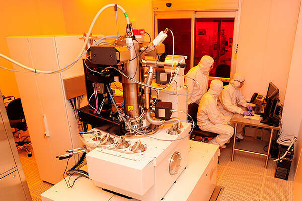 Three people in clean room uniforms at monitor next to e beam lithography system