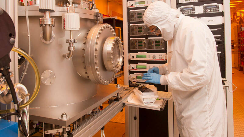 A person in clean room suit inspects material wafers outside a dual-chamber sputtering machine